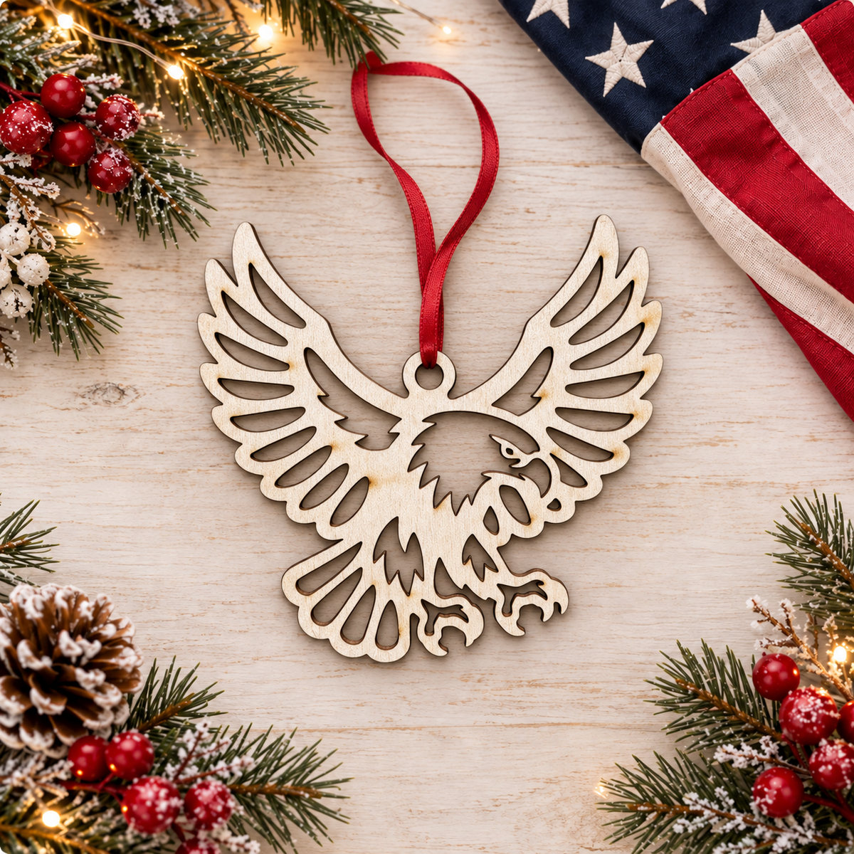 A laser-cut wooden eagle Christmas ornament with a red hanging ribbon, displayed on a light wood surface surrounded by festive pine branches, red berries, a pinecone, and a folded American flag.