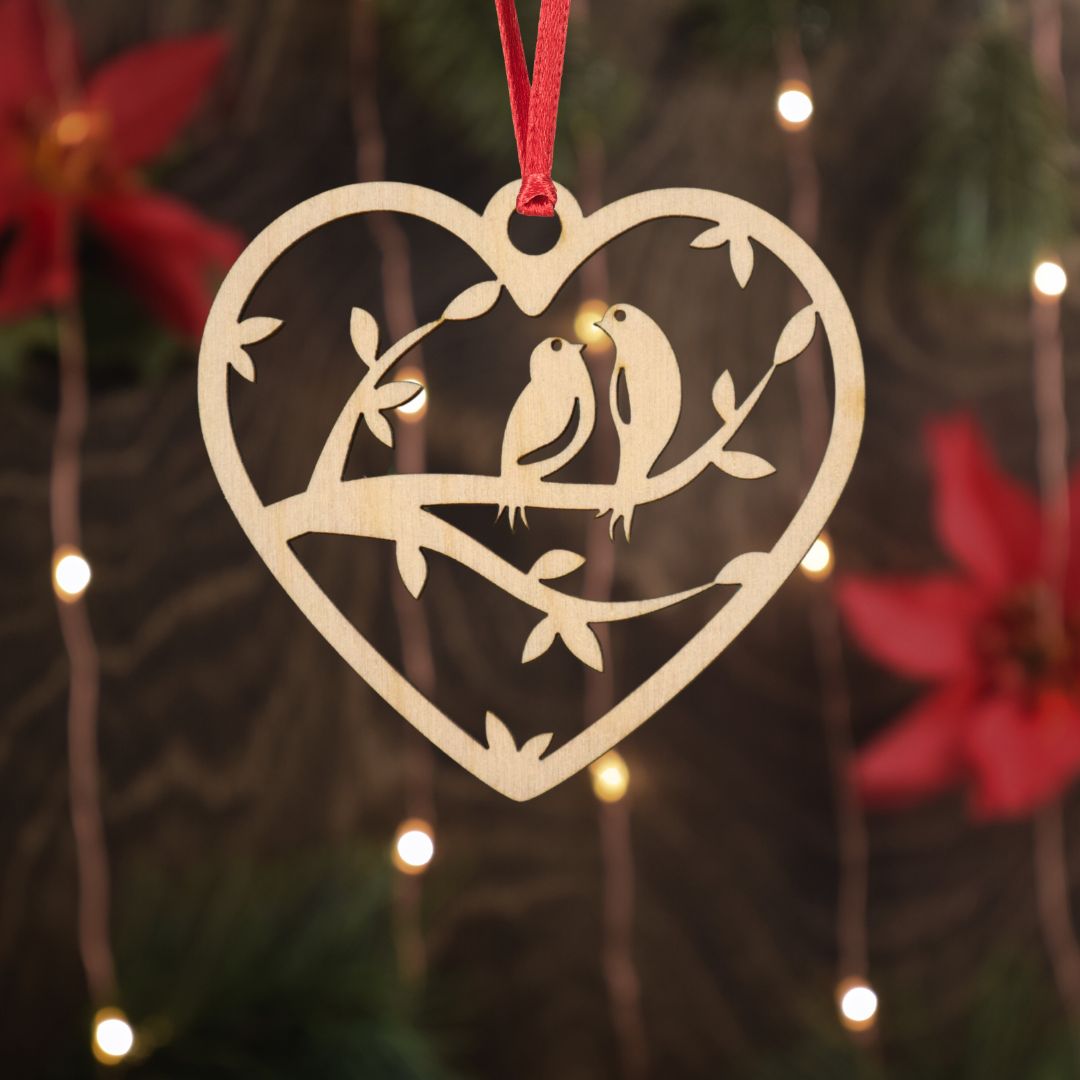 Heart-shaped ornament with cut-out birds on a branch against a festive background with lights and flowers.