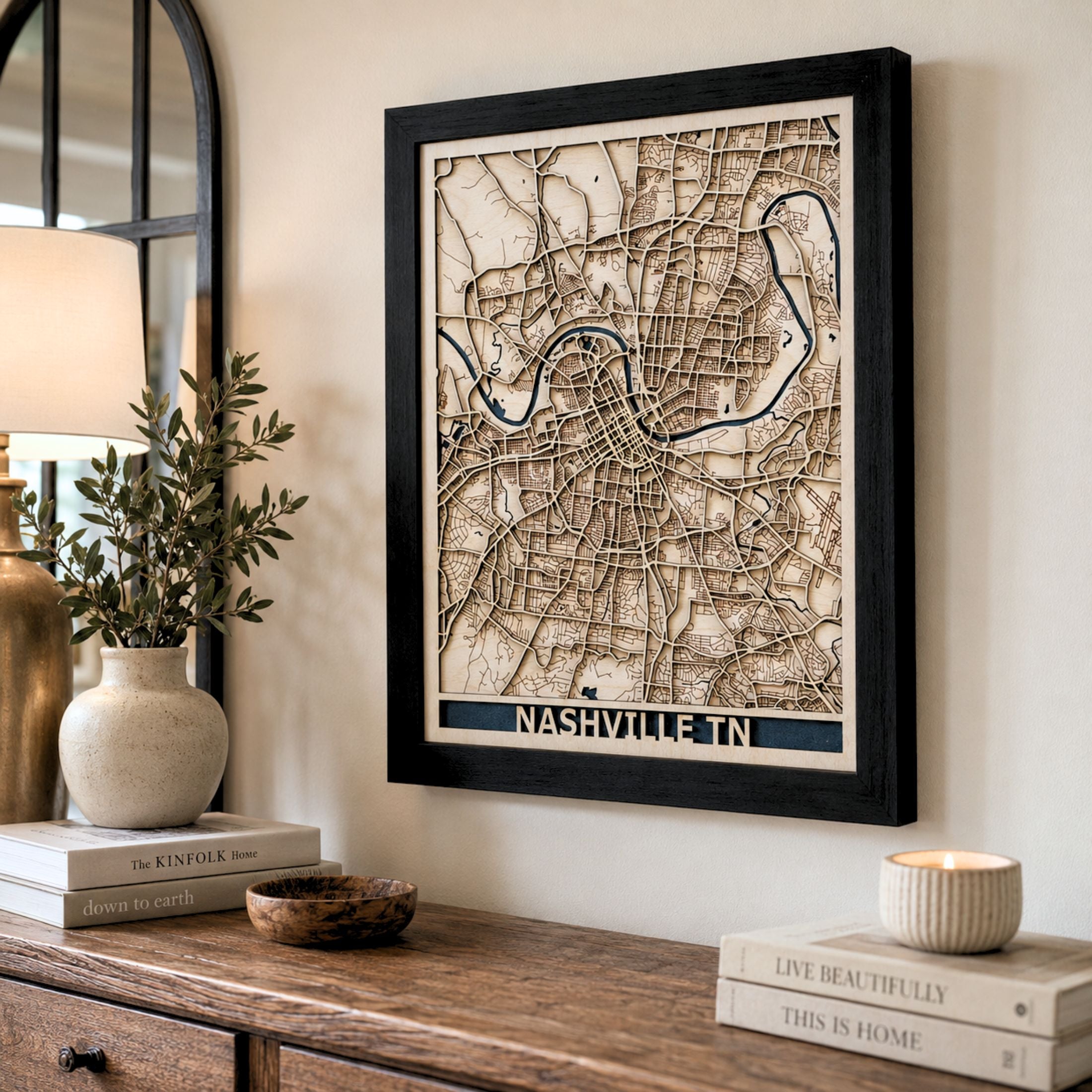 Framed wooden Nashville city map wall art hanging on a beige wall above a wooden console table, styled with books, a vase, and a candle.