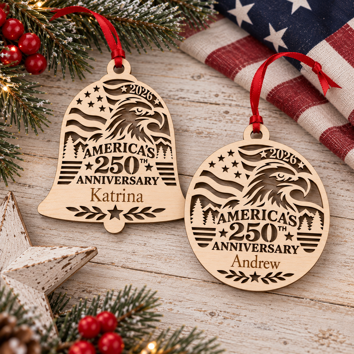 Two personalized wooden Christmas ornaments shaped like a bell and a round disc, both engraved with an eagle, stars, stripes, and the text 'America's 250th Anniversary 2026' and names 'Katrina' and 'Andrew', hanging from red ribbons on a rustic wood surface with evergreen branches, red berries, a star, and an American flag in the background.