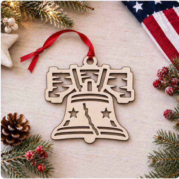 A laser-cut wooden Liberty Bell Christmas ornament with a red ribbon hanger, displayed on a light wood surface surrounded by festive holiday decorations including pine branches, a pinecone, red berries, and a corner of an American flag.