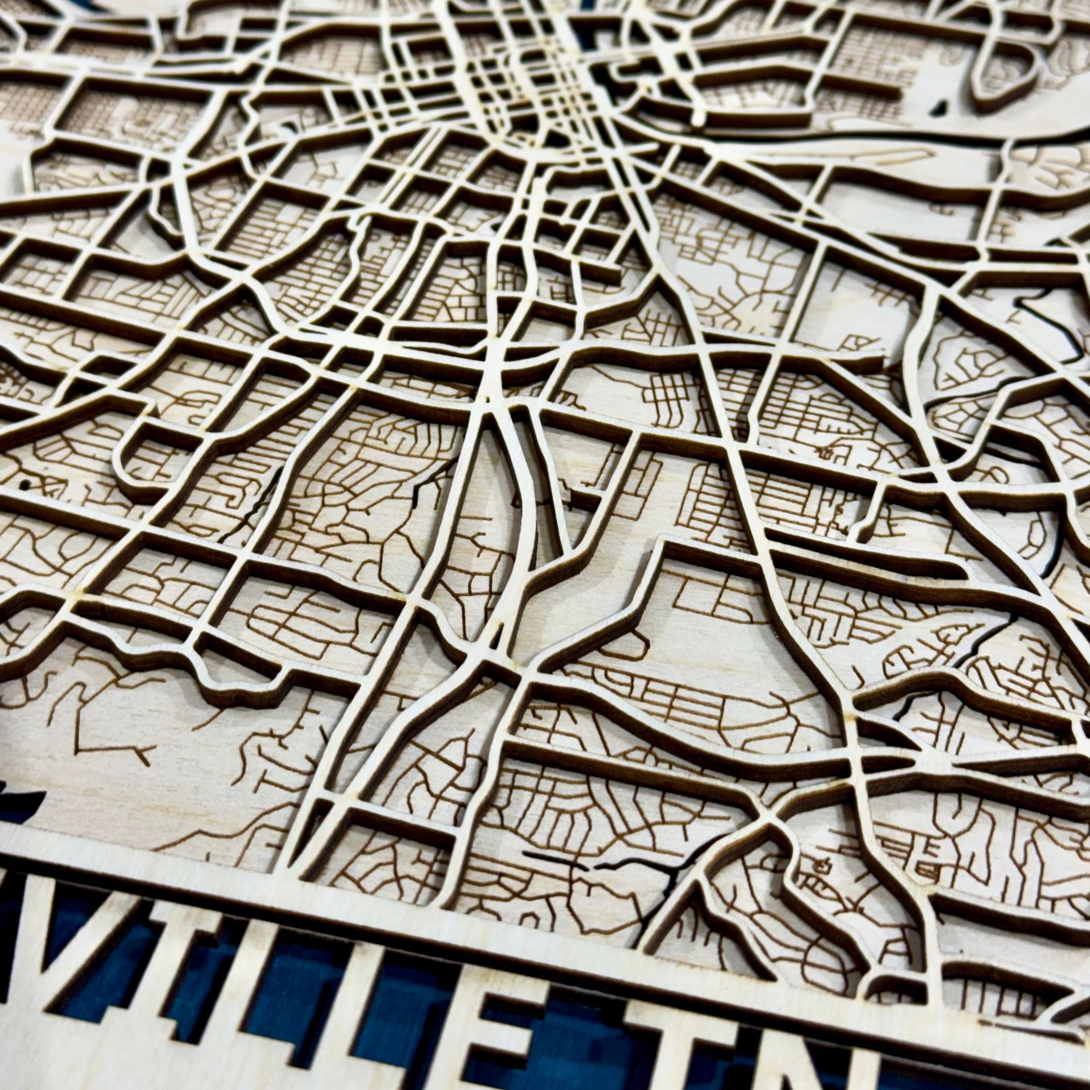 Close-up of a laser-engraved wooden city map, showing detailed street lines and labels with a natural wood finish and a dark blue background visible in the lower section.