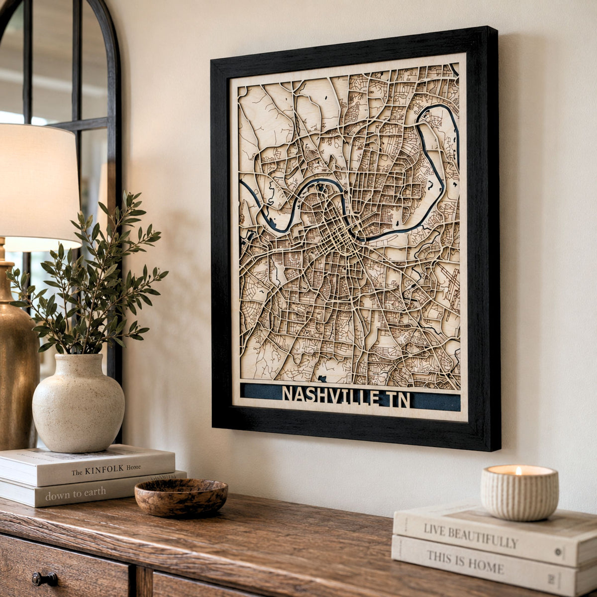 Framed wooden Nashville city map wall art hanging on a beige wall above a wooden console table, styled with books, a vase, and a candle.