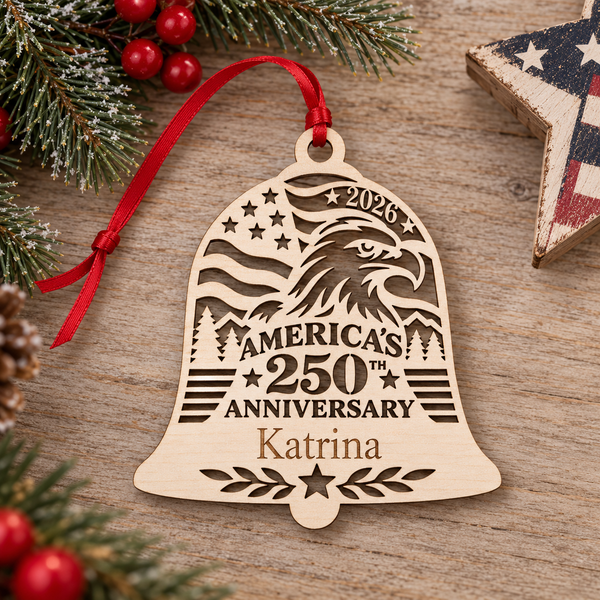 A personalized wooden Christmas bell ornament engraved with an eagle, stars, and the text '2026 America's 250th Anniversary Katrina,' hanging from a red ribbon on a rustic wood surface with holiday greenery and red berries around it.