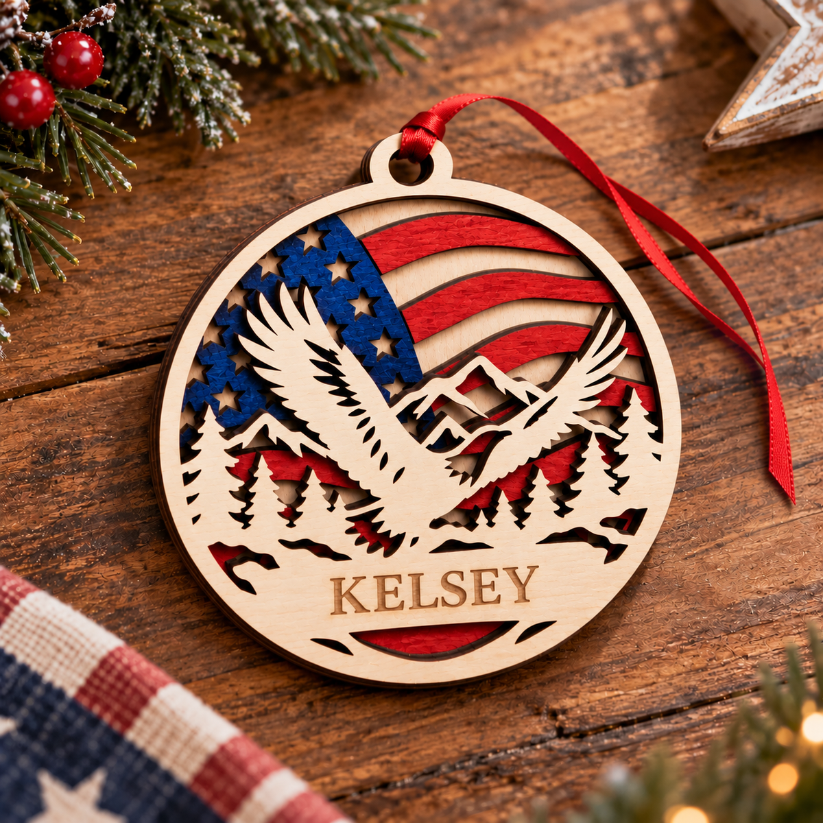 Personalized round wooden Christmas ornament featuring a laser-cut eagle, mountains, pine trees, and an American flag design, with the name 'Kelsey' engraved at the bottom and a red ribbon hanging from it, displayed on a rustic wooden surface with holiday decorations.