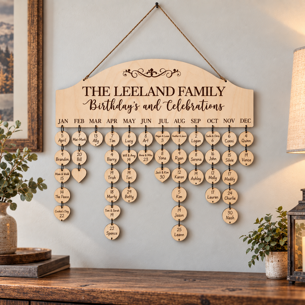 A personalized wooden family birthday and celebration wall chart hanging on a light gray wall above a wooden console table. The sign reads 'THE LEELAND FAMILY Birthday's and Celebrations' and features rows of small round wooden discs labeled with months and names, plus hanging heart-shaped plaques.