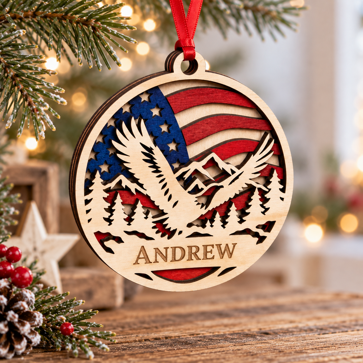 Personalized wooden Christmas ornament featuring a laser-cut eagle, mountains, pine trees, and an American flag design, with the name ANDREW engraved at the bottom and hanging from a red ribbon.