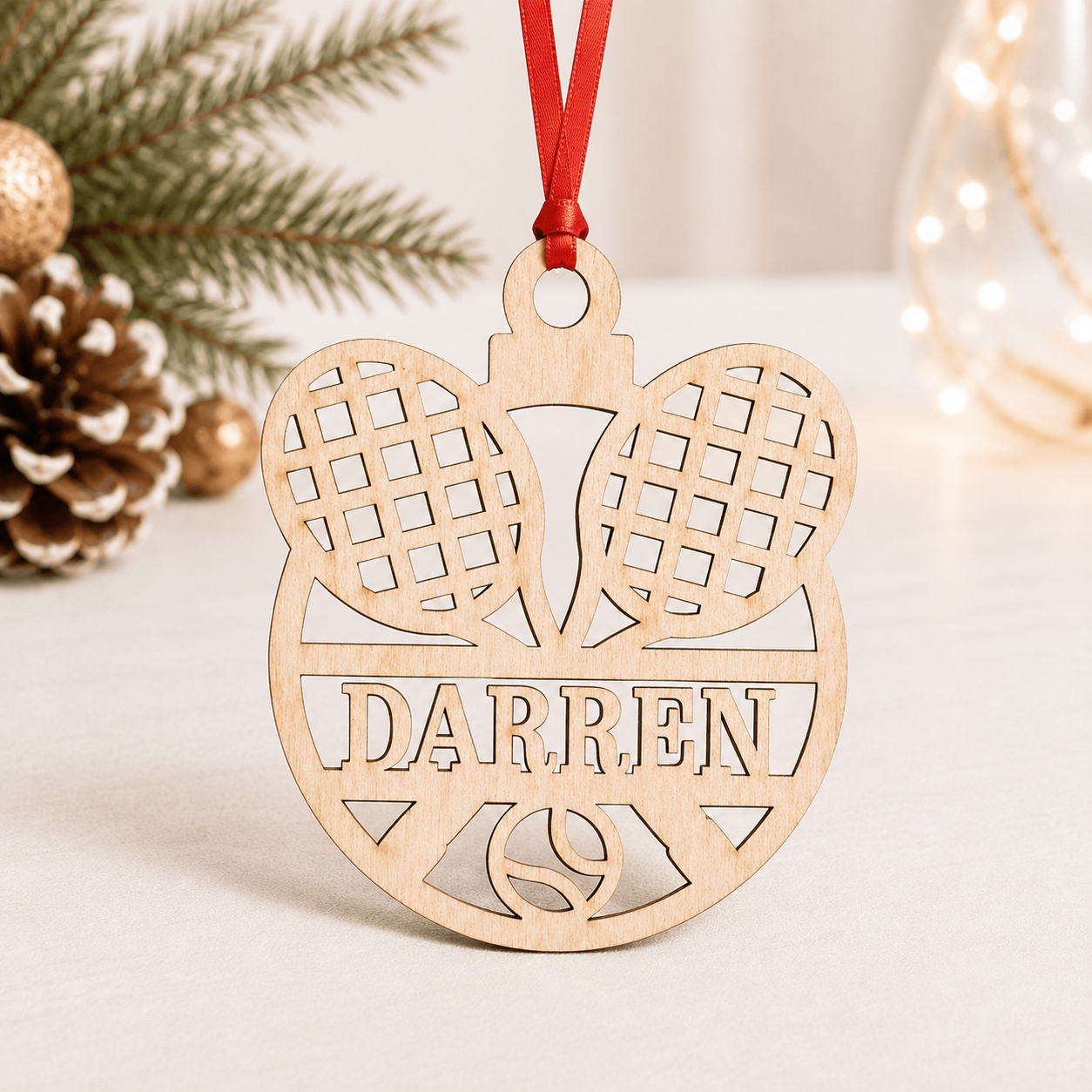 A personalized wooden Christmas ornament shaped like a tennis player's head silhouette with two tennis rackets, featuring the name 'DARREN' and a red hanging ribbon, styled on a light tabletop with pine branches and a pinecone in the background.